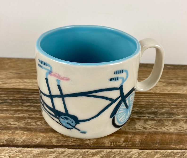 K&ouml;nnte beinhalten: Cremefarbene Keramiktasse mit blauem Innenraum und einem Fahrradmuster in Marineblau und Hellblau. Die Fahrradillustration umschlie&szlig;t die Tasse, mit rosa Akzenten am Lenker. Die Tasse hat einen gebogenen Griff.