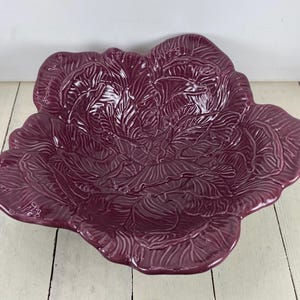 May include: A large, burgundy-colored ceramic serving bowl shaped like a cabbage. The bowl features detailed embossed leaf patterns and a glossy finish. The bowl is placed on a white wooden surface.