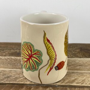 K&ouml;nnte beinhalten: Eine cremefarbene Keramiktasse mit farbenfrohen botanischen Illustrationen. Das Design umfasst ein gro&szlig;es gr&uuml;nes und rotes Blatt, ein gelbes und rotes Blatt und einen braunen Stiel. Die Tasse hat einen einfachen Henkel und eine glatte, gl&auml;nzende Oberfl&auml;che.