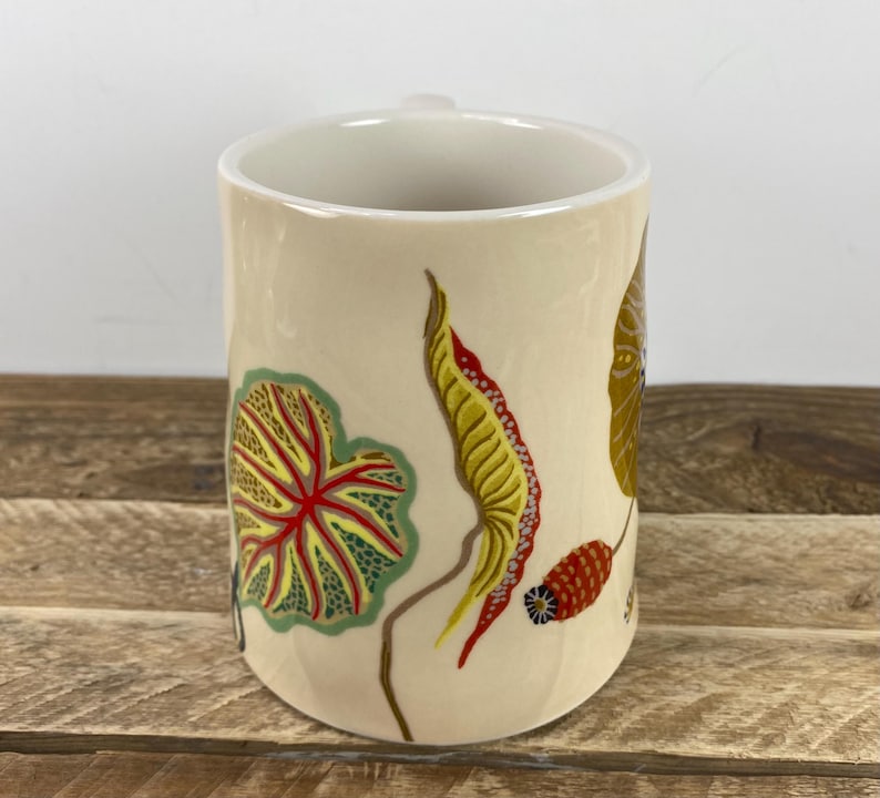 K&ouml;nnte beinhalten: Eine cremefarbene Keramiktasse mit farbenfrohen botanischen Illustrationen. Das Design umfasst ein gro&szlig;es gr&uuml;nes und rotes Blatt, ein gelbes und rotes Blatt und einen braunen Stiel. Die Tasse hat einen einfachen Henkel und eine glatte, gl&auml;nzende Oberfl&auml;che.