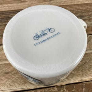 K&ouml;nnte beinhalten: Wei&szlig;er Keramikbecher mit einer blauen Fahrradillustration und dem Wort "ANTHROPOLOGIE" auf der Unterseite. Der Becher hat einen Henkel und steht auf einer Holzoberfl&auml;che.