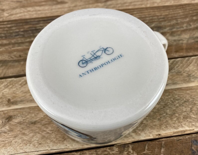 K&ouml;nnte beinhalten: Wei&szlig;er Keramikbecher mit einer blauen Fahrradillustration und dem Wort "ANTHROPOLOGIE" auf der Unterseite. Der Becher hat einen Henkel und steht auf einer Holzoberfl&auml;che.