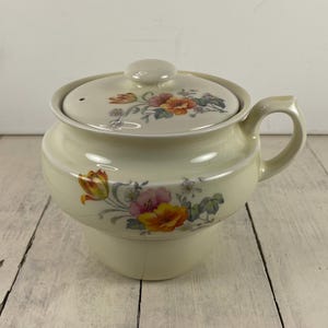 May include: Cream-colored ceramic teapot with a lid and handle. The teapot is decorated with floral patterns in shades of orange, pink, and yellow. The lid has a small knob and a vent hole. The teapot is sitting on a light-colored wooden surface.