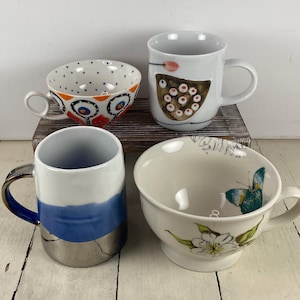 K&ouml;nnte beinhalten: Vier Keramikbecher und -schalen mit einzigartigen Designs. Eine Tasse hat ein blau-wei&szlig;es Muster, eine andere ein Schmetterlings- und Blumendesign, und eine dritte ein braun-wei&szlig;es Design. Die vierte Tasse hat ein blau-silbernes Design.