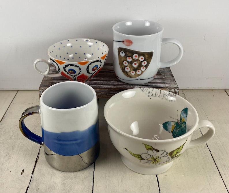 K&ouml;nnte beinhalten: Vier Keramikbecher und -schalen mit einzigartigen Designs. Eine Tasse hat ein blau-wei&szlig;es Muster, eine andere ein Schmetterlings- und Blumendesign, und eine dritte ein braun-wei&szlig;es Design. Die vierte Tasse hat ein blau-silbernes Design.