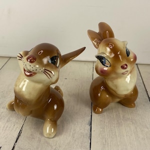 May include: Two vintage ceramic bunny figurines. One bunny is posed with an ear up and a smiling face, the other bunny has its ears up and is looking to the side. Both bunnies have brown fur, cream-colored faces, and rosy cheeks.