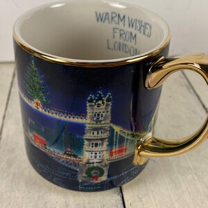 K&ouml;nnte beinhalten: Eine Keramik-Tasse mit goldenem Henkel und Rand, die eine n&auml;chtliche Londoner Szene mit einem Weihnachtsbaum und der Tower Bridge zeigt. Der Text "Warm Wishes From London" steht im Inneren.