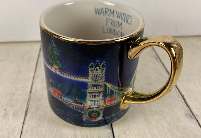 K&ouml;nnte beinhalten: Eine Keramik-Tasse mit goldenem Henkel und Rand, die eine n&auml;chtliche Londoner Szene mit einem Weihnachtsbaum und der Tower Bridge zeigt. Der Text "Warm Wishes From London" steht im Inneren.