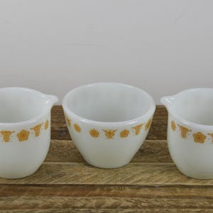May include: Three white vintage milk glass serving pieces. Two pitchers with handles and spouts, and a small bowl. Each piece has a gold floral pattern around the rim. The set is displayed on a wooden surface.
