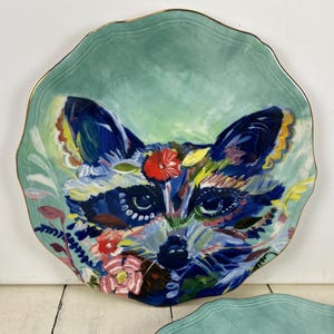 May include: A decorative ceramic plate with a raccoon face design. The plate features a teal background with a gold rim. The raccoon's face is painted with vibrant colors, including blue, red, and yellow, and is surrounded by floral accents.
