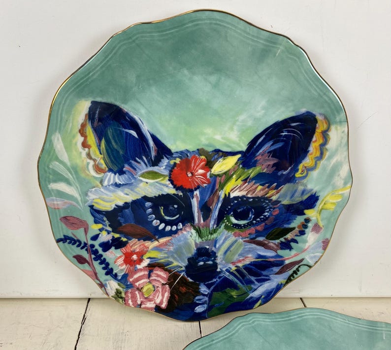 May include: A decorative ceramic plate with a raccoon face design. The plate features a teal background with a gold rim. The raccoon's face is painted with vibrant colors, including blue, red, and yellow, and is surrounded by floral accents.