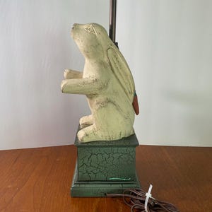 May include: A table lamp featuring a cream-colored rabbit figurine with a distressed finish, standing on a rectangular base with a dark green crackle paint effect. A brown electrical cord with a white plastic tie extends from the base.
