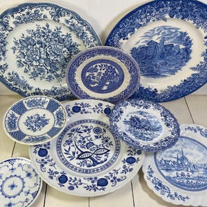 May include: A collection of eight white ceramic plates featuring blue transferware patterns. The designs include floral arrangements, a landscape with a building, a horse-drawn carriage, a windmill scene, and intricate geometric borders.
