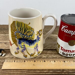 K&ouml;nnte beinhalten: Eine cremefarbene Keramiktasse mit einer skurrilen Illustration eines Fabelwesens. Eine Dose Campbell's Tomatensuppe und eine kleine Corgi-Figur befinden sich neben der Tasse. Ein Holzlineal liegt im Vordergrund.