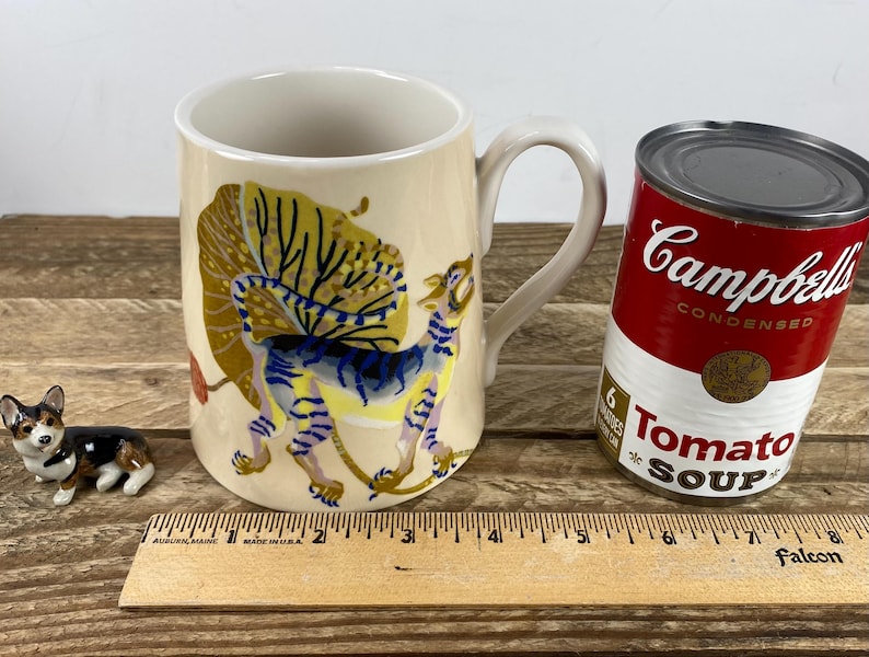 K&ouml;nnte beinhalten: Eine cremefarbene Keramiktasse mit einer skurrilen Illustration eines Fabelwesens. Eine Dose Campbell's Tomatensuppe und eine kleine Corgi-Figur befinden sich neben der Tasse. Ein Holzlineal liegt im Vordergrund.
