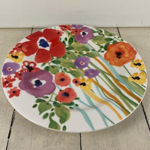May include: A white ceramic plate with a colorful floral design. The plate features red, purple, orange, and yellow flowers with green leaves. The flowers are painted in a watercolor style, with a blue center. The plate is round and has a slightly raised edge.