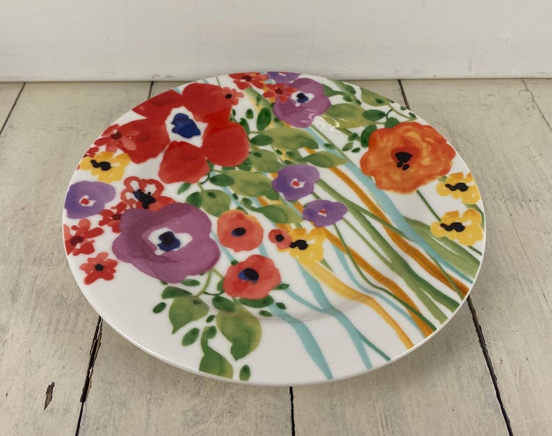 May include: A white ceramic plate with a colorful floral design. The plate features red, purple, orange, and yellow flowers with green leaves. The flowers are painted in a watercolor style, with a blue center. The plate is round and has a slightly raised edge.
