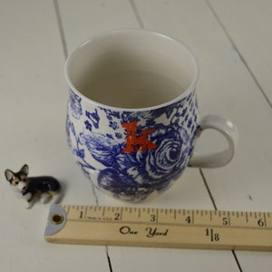 May include: A white ceramic mug with a blue floral pattern and a red letter "K". A small corgi figurine and a wooden ruler are next to the mug. The ruler measures up to 8 inches.