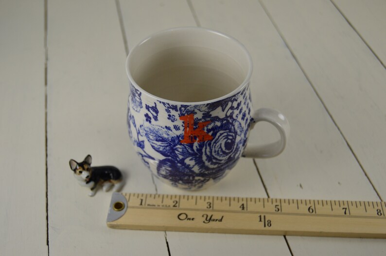 May include: A white ceramic mug with a blue floral pattern and a red letter "K". A small corgi figurine and a wooden ruler are next to the mug. The ruler measures up to 8 inches.