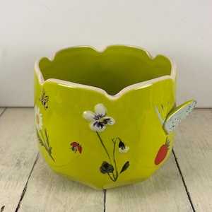 Może przedstawiać: Chartreuse ceramiczna donica z falistym brzegiem i uchwytem w kształcie motyla. Donica jest ozdobiona malowanymi kwiatami, biedronką i motylem. Wnętrze jest w kolorze jasnozielonym.