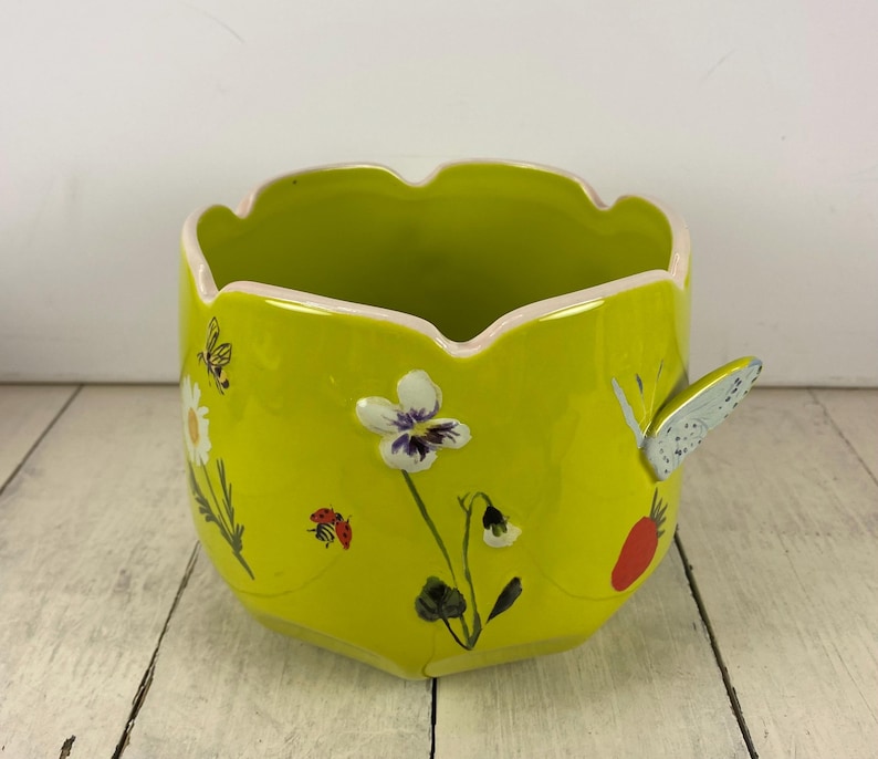 Może przedstawiać: Chartreuse ceramiczna donica z falistym brzegiem i uchwytem w kształcie motyla. Donica jest ozdobiona malowanymi kwiatami, biedronką i motylem. Wnętrze jest w kolorze jasnozielonym.