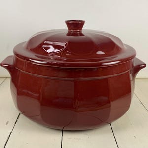May include: A deep, glossy, dark red ceramic casserole dish with a matching lid. The pot features a rounded, scalloped body with two integrated side handles and a central knob on the lid. The surface has a smooth, reflective finish throughout.