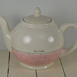 May include: White ceramic teapot with a pink and white polka dot design. The teapot has a lid and a handle. The text "TEA TIME." is printed on the teapot.