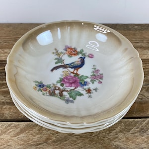 May include: A stack of four small, scalloped-edge porcelain dishes. Each dish features a central design of a blue bird perched on a flowering branch with pink, orange, and blue blossoms. The rims have a light brown, marbled effect.