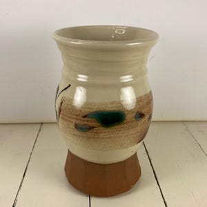 May include: A ceramic vase with a wide, flared rim and a rounded body. The upper portion is cream-colored with a horizontal band of brown speckled glaze featuring small teal accents. The base is a solid, unglazed terracotta-colored clay.