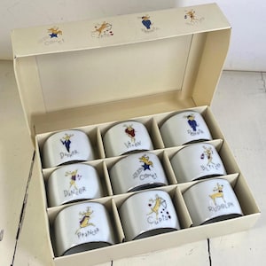 May include: A set of nine white ceramic napkin rings housed in a cream-colored cardboard box. Each ring features a unique illustration of a reindeer with a name printed below, including Dasher, Dancer, Prancer, Vixen, Comet, Cupid, Donner, Blitzen, and Rudolph.