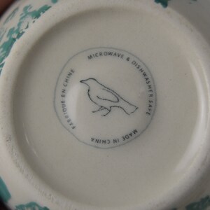 May include: White ceramic bowl with a green floral design. The bottom of the bowl has a black and white label that reads "Microwave & Dishwasher Safe. Made in China. Fabrique en Chine."