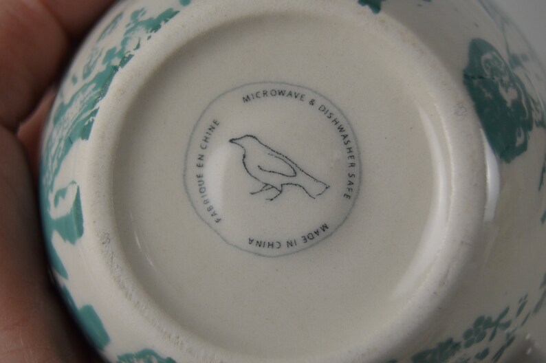 May include: White ceramic bowl with a green floral design. The bottom of the bowl has a black and white label that reads "Microwave & Dishwasher Safe. Made in China. Fabrique en Chine."