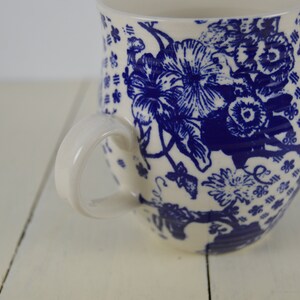 May include: White ceramic mug with a blue floral pattern. The mug features a large handle and a detailed design of flowers and leaves. The background is a white wooden surface.