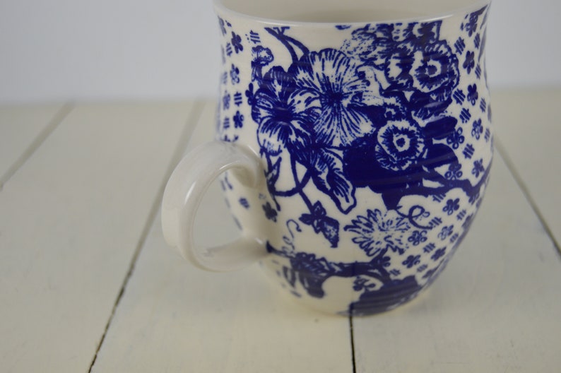 May include: White ceramic mug with a blue floral pattern. The mug features a large handle and a detailed design of flowers and leaves. The background is a white wooden surface.
