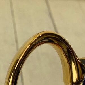 K&ouml;nnte beinhalten: Nahaufnahme eines goldfarbenen Griffs, m&ouml;glicherweise von einer Teetasse oder einem Becher. Der Griff hat ein glattes, geschwungenes Design mit gl&auml;nzendem Finish. Der Griff ist an einer dunkelblauen Oberfl&auml;che befestigt.