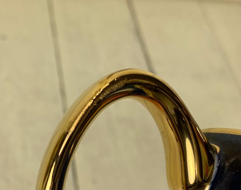 K&ouml;nnte beinhalten: Nahaufnahme eines goldfarbenen Griffs, m&ouml;glicherweise von einer Teetasse oder einem Becher. Der Griff hat ein glattes, geschwungenes Design mit gl&auml;nzendem Finish. Der Griff ist an einer dunkelblauen Oberfl&auml;che befestigt.