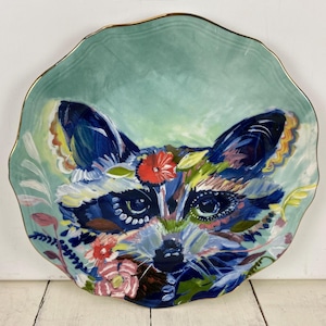 May include: A decorative ceramic plate with a hand-painted raccoon design. The plate features a teal background with a gold rim. The raccoon is depicted with blue, white, red, and yellow floral accents.