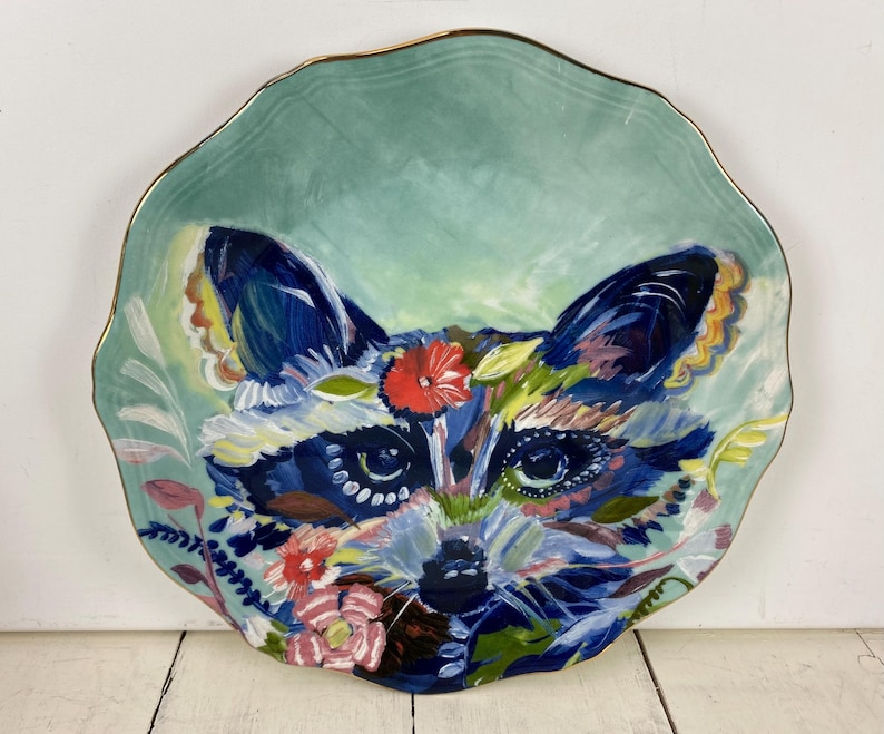 May include: A decorative ceramic plate with a hand-painted raccoon design. The plate features a teal background with a gold rim. The raccoon is depicted with blue, white, red, and yellow floral accents.