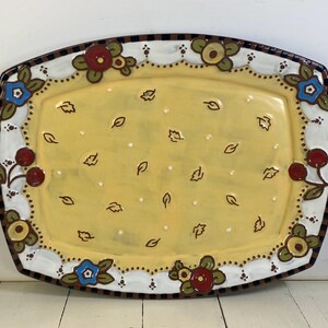 May include: An oval ceramic serving platter with a yellow center featuring small brown leaf motifs and white dots. The white border is decorated with red, blue, and yellow floral patterns and cherries, finished with a black and brown checkered rim.