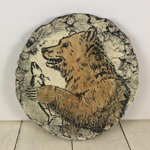 May include: A round, rustic ceramic plate featuring a brown bear holding a fish. The bear is depicted in a detailed, stylized illustration with black outlines and a beige background. The plate has a textured, cracked appearance.