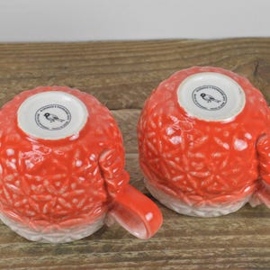 May include: Two red ceramic mugs with a white floral design and a bird logo on the bottom. The mugs have a white rim and a white base.