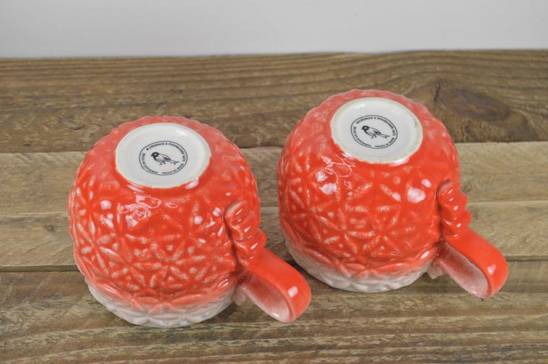 May include: Two red ceramic mugs with a white floral design and a bird logo on the bottom. The mugs have a white rim and a white base.