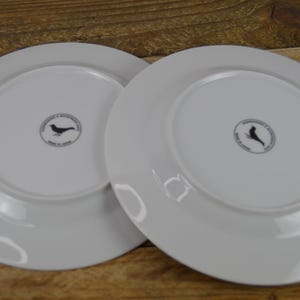 Puede incluir: Dos platos de cena de cer&aacute;mica blanca con un dise&ntilde;o de p&aacute;jaro negro y el texto "Hecho a mano en Espa&ntilde;a" en la parte inferior.