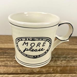 Puede incluir: Taza de cer&aacute;mica color crema con borde y asa negros. La taza presenta las palabras "MORE please" en un dise&ntilde;o ovalado negro. La taza est&aacute; sobre una superficie de madera.