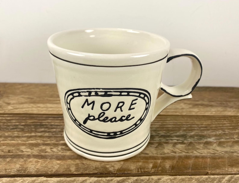 Puede incluir: Taza de cer&aacute;mica color crema con borde y asa negros. La taza presenta las palabras "MORE please" en un dise&ntilde;o ovalado negro. La taza est&aacute; sobre una superficie de madera.