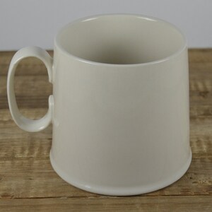 Pode incluir: Uma caneca de cer&acirc;mica branca com uma al&ccedil;a arredondada. A caneca est&aacute; sobre uma superf&iacute;cie de madeira.