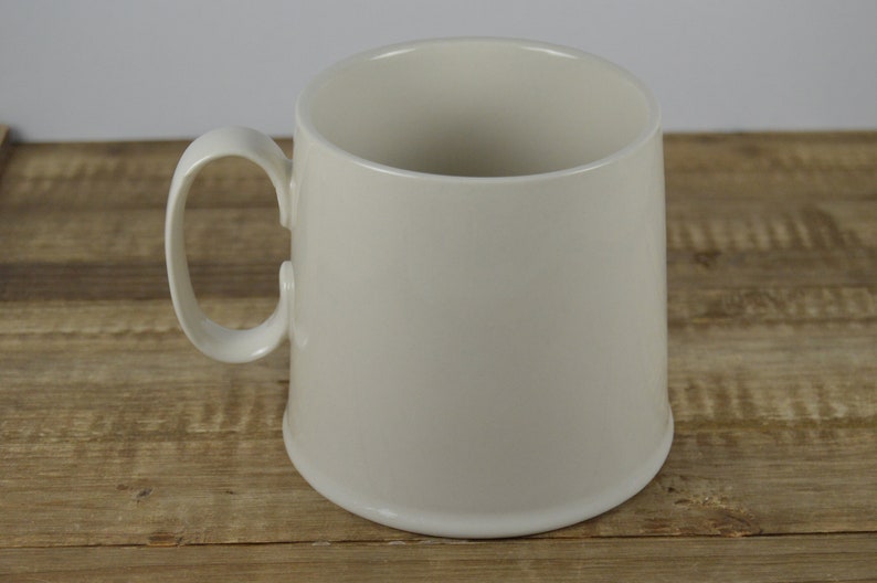 Pode incluir: Uma caneca de cer&acirc;mica branca com uma al&ccedil;a arredondada. A caneca est&aacute; sobre uma superf&iacute;cie de madeira.