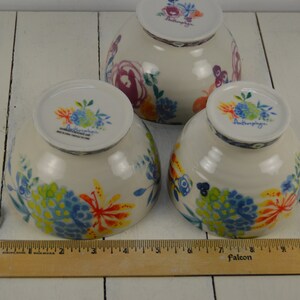 May include: Three white ceramic bowls with a floral pattern in shades of blue, yellow, orange, and green. The bowls are all the same size and have a handle on the side. The bowls are sitting on a white wooden surface. The text "Anthropologie" is visible on the bottom of each bowl.
