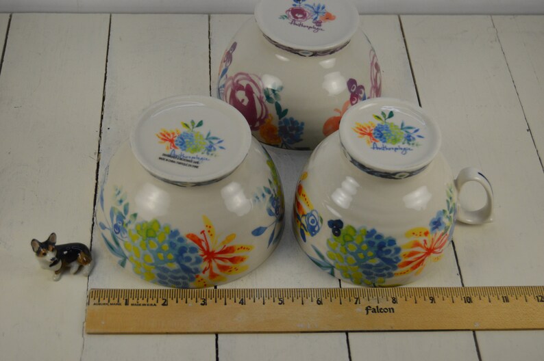 May include: Three white ceramic bowls with a floral pattern in shades of blue, yellow, orange, and green. The bowls are all the same size and have a handle on the side. The bowls are sitting on a white wooden surface. The text "Anthropologie" is visible on the bottom of each bowl.