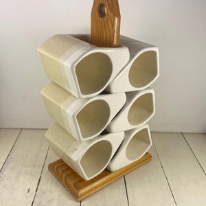 May include: A set of six off-white ceramic mugs with a speckled texture, arranged in two vertical columns on a wooden stand. The mugs feature a distinct angular, geometric shape and are held in place by a central wooden support and a rectangular base.
